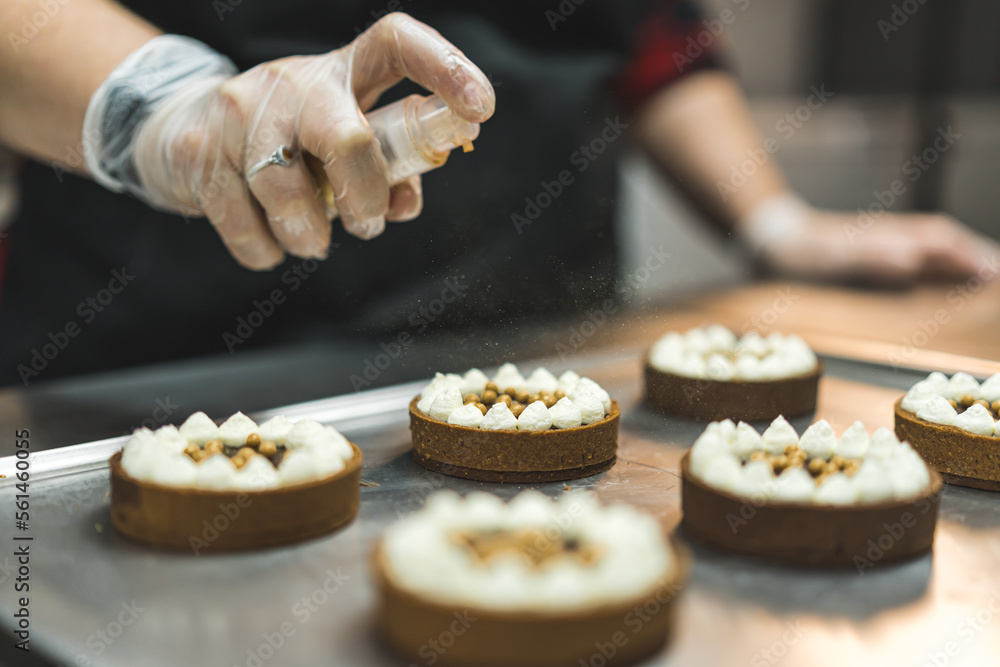 Professional female baker wearing gloves and black apron spraying golden edible glitter onto mini dessert chocolate tarts. Baking process. Decorating. Horizontal indoor close-up shot. High quality