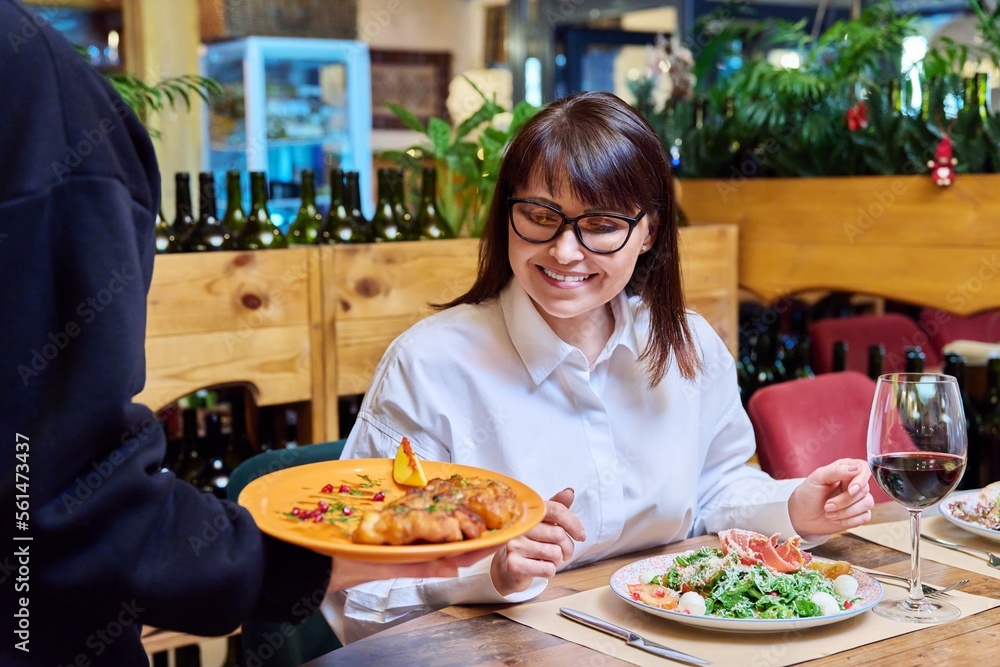 Woman sitting in restaurant, rejoices at plate of cooked food in hands of waiter