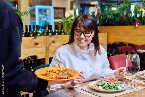 Woman sitting in restaurant, rejoices at plate of cooked food in hands of waiter