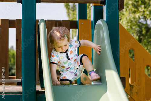 Happy blond little toddler girl having fun and sliding on outdoor playground. Positive funny baby child smiling. Summer leisure for small children, active games for family outdoors. © Irina Schmidt