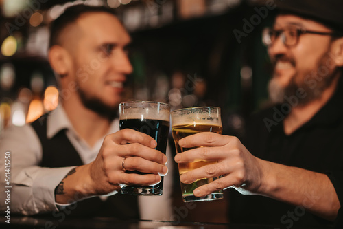 two happy smiling male friends clink glasses of draft beer at bar counter in pub