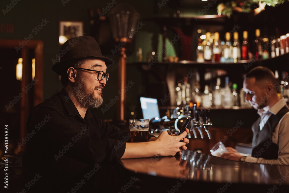 happy smiling man uses a mobile phone smartphone while sitting at bar counter with a beer in pub