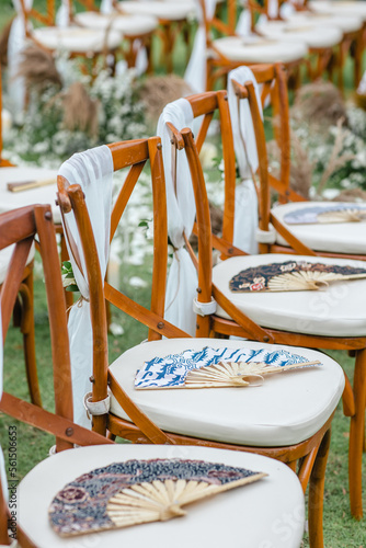 Wedding wooden chairs decorated with asian fan called Shan. Rustic aisle chairs standing on lawn for ceremony in garden with wooden and fabric hand fan on each. photo