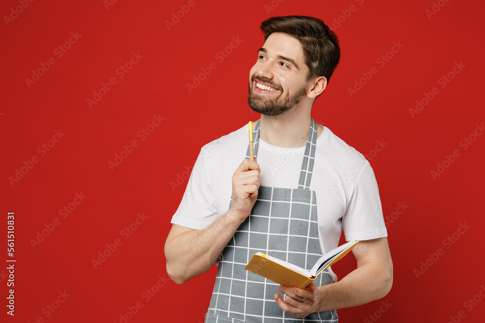 Young minded waiter fun happy male housewife housekeeper chef cook baker man wears grey apron hold write in notebook look aside on area isolated on plain red background studio. Cooking food concept. Young minded waiter fun happy male housewife housekeeper chef cook baker man wears grey apron hold write in notebook look aside on area isolated on plain red background studio. Cooking food concept.