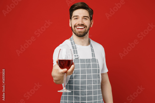 Young smiling sommelier happy male housewife housekeeper chef cook baker man wear grey apron hold in hand give glass of wine look camera isolated on plain red background studio. Cooking food concept.
