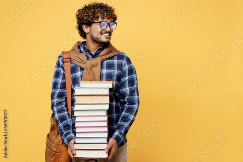 Obraz na plátně Young teen Indian boy IT student wear casual clothes shirt glasses bag hold in hands pile of books look aside on area isolated on plain yellow color background High school university college concept