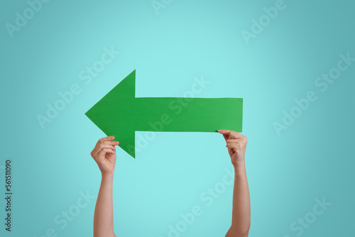 Close up of  female hand holding big green paper arrow, isolated over blue studio background wall with copy space for advertisement. left direction. Advertising area, mockup