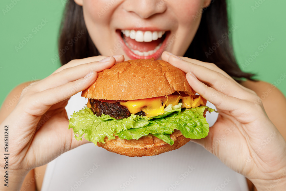 Cropped close up young happy woman wear white clothes holding eating biting tasty burger isolated on plain pastel light green background. Proper nutrition healthy fast food unhealthy choice concept.