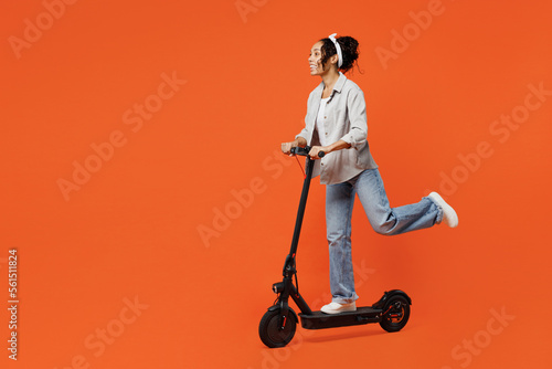 Full body young happy fun woman of African American ethnicity she wears grey shirt headband riding e-scooter look aside isolated on plain orange background studio portrait. People lifestyle concept.