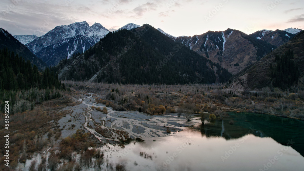 Issyk mountain lake with mirror water at sunset. The color of the water changes before our eyes. There are trees in clear water. Snowy mountains and green hills are visible. Clouds are reflected Issyk mountain lake with mirror water at sunset. The color of the water changes before our eyes. There are trees in clear water. Snowy mountains and green hills are visible. Clouds are reflected