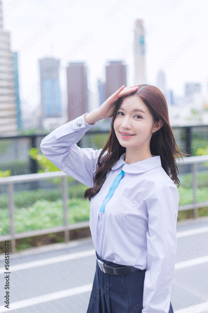 Cute Asian high school student girl in school uniform with braces on her teeth stands she raised her hand to block sunlight and smiles confidently while she looks at the camera happily with the city