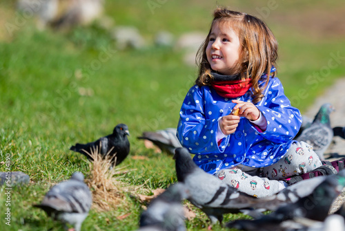 Caucasian blonde little girl feeding pigeons in a park on a sunny day. Girl sitting on the ground next to some pigeons. Girls and animals.