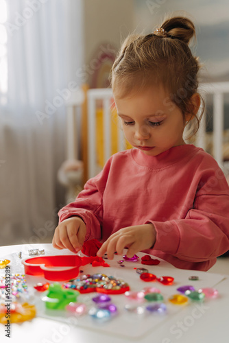 A little girl playing with red heart from play dough for modeling and decorations from crystal rhinestones and shiny stones. Toddlers crafts for Valentine's Day. Holiday Art Activity for Kids.