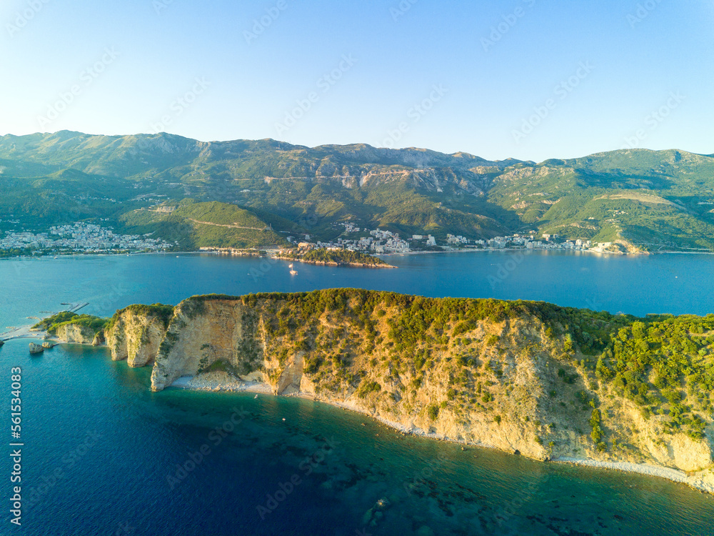 Island of St. Nicholas with vegetation on shores in the Adriatic Sea against the backdrop of coastal towns, the Montenegrin mountains and clear sky