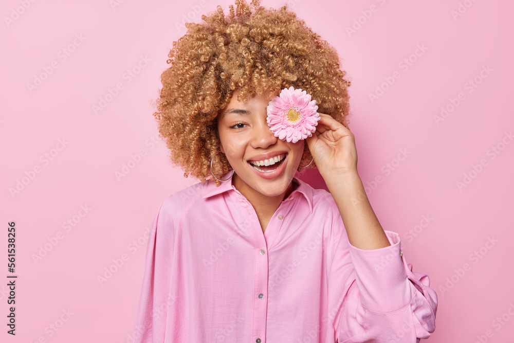 Positive young woman with curly hair covers eye with gerbera flower smiles broadly shows white teeth dressed in shirt isolated over pink background enjoys pleasant fragrance has romantic mood