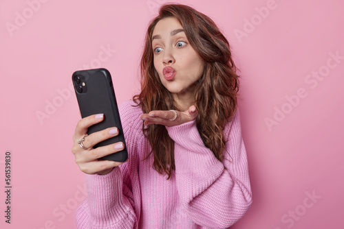 Romantic beautiful dark haired young woman blows air kiss at camera has video chat with boyfriend expresses love keeps lips rounded dressed in loose knitted jumper isolated over pink background
