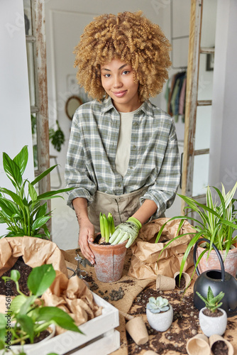 Vertical shot of curly haired woman busy transplanting potted flowers surrounded by soil and gardening tools wears checkered shirt and glove stands indoor against home interior. House plants care