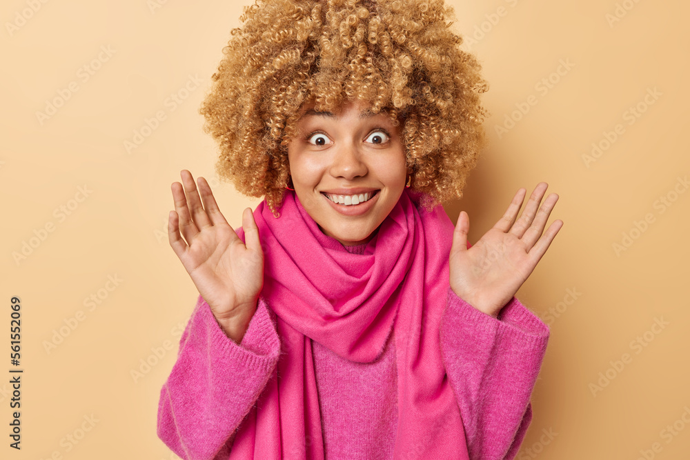 Horizontal shot of cheerful curly haired woman reacts to amazing news keeps palms raised dressed in pink jumper and scarf around neck hears something awesome isolated over beige studio background Horizontal shot of cheerful curly haired woman reacts to amazing news keeps palms raised dressed in pink jumper and scarf around neck hears something awesome isolated over beige studio background
