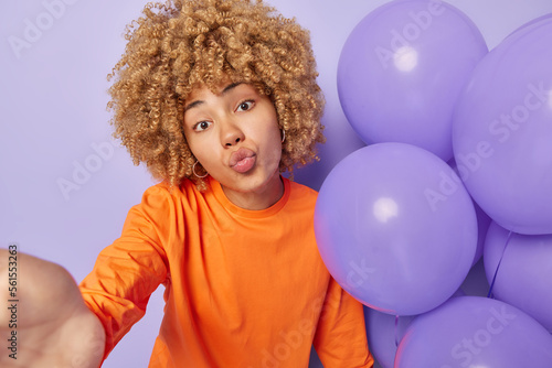 Romantic woman makes selfie poses near inflated balloons keeps lips folded sends air kiss at camera dressed in orange sweater celebrates anniversary isolated on purple background. People and holidays