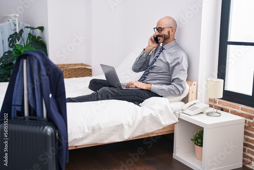 Young hispanic man business worker using laptop talking on smartphone at hotel room