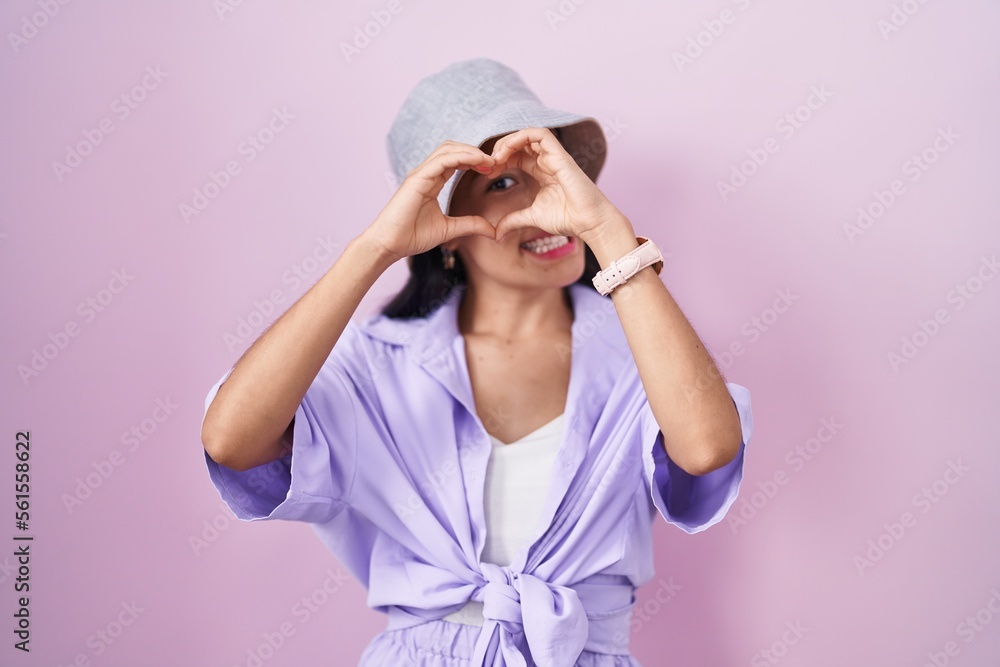 Young hispanic woman standing over pink background wearing hat doing heart shape with hand and fingers smiling looking through sign Young hispanic woman standing over pink background wearing hat doing heart shape with hand and fingers smiling looking through sign