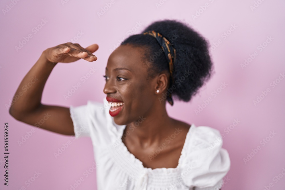 African woman with curly hair standing over pink background very happy and smiling looking far away with hand over head. searching concept. African woman with curly hair standing over pink background very happy and smiling looking far away with hand over head. searching concept.