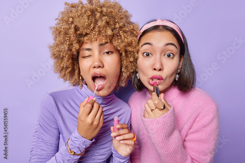 Photo of young multiracial women apply lipstick and lipgloss want to look beautiful get prepared for party or date dressed in casual jumpers isolated over purple background. Beauty procedures photo