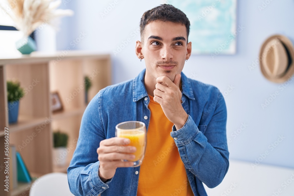 Handsome hispanic man drinking glass of orange juice serious face thinking about question with hand on chin, thoughtful about confusing idea Handsome hispanic man drinking glass of orange juice serious face thinking about question with hand on chin, thoughtful about confusing idea
