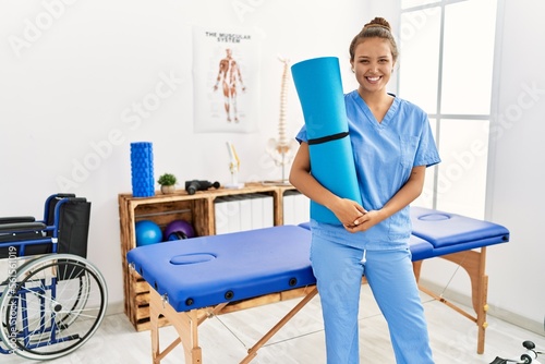 Young beautiful hispanic woman physiotherapist smiling confident holding foam roller at rehab clinic