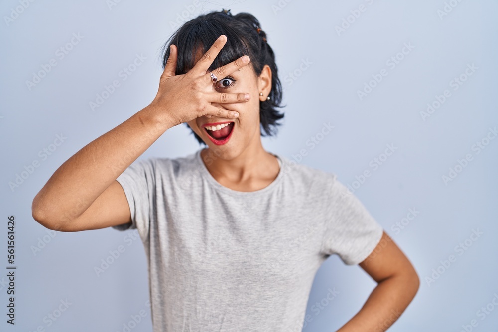 Young hispanic woman wearing casual t shirt over blue background peeking in shock covering face and eyes with hand, looking through fingers with embarrassed expression. Young hispanic woman wearing casual t shirt over blue background peeking in shock covering face and eyes with hand, looking through fingers with embarrassed expression.