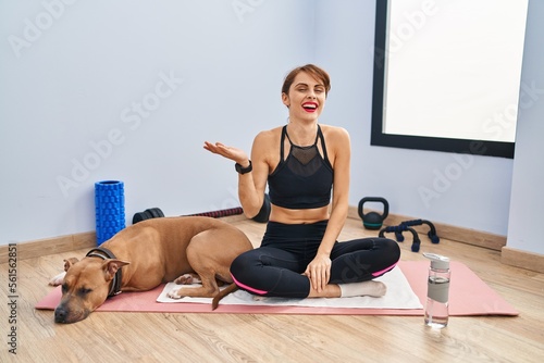 Young beautiful woman sitting on yoga mat smiling cheerful presenting and pointing with palm of hand looking at the camera.