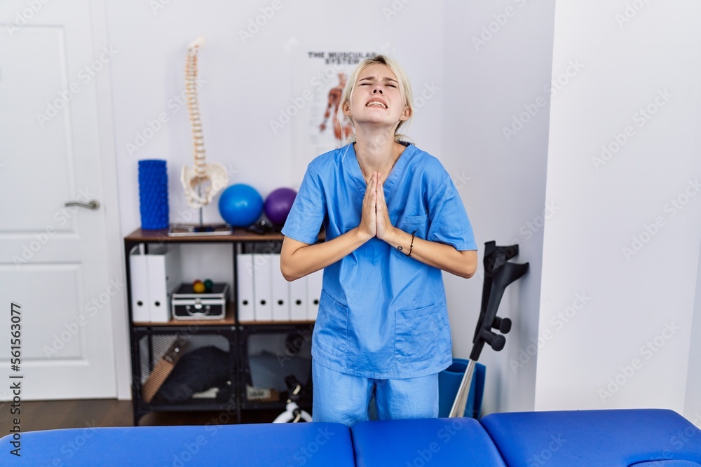 Young physiotherapist woman working at pain recovery clinic begging and praying with hands together with hope expression on face very emotional and worried. begging. Young physiotherapist woman working at pain recovery clinic begging and praying with hands together with hope expression on face very emotional and worried. begging.