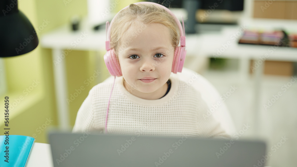 Adorable blonde girl student using laptop and headphones sitting on table at classroom Adorable blonde girl student using laptop and headphones sitting on table at classroom