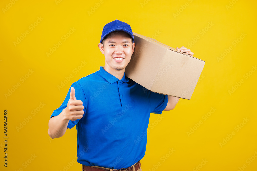 Portrait excited delivery happy man logistic standing he smile wearing blue t-shirt and cap uniform holding parcel box show thumb up finger looking to camera, studio shot isolated on yellow background Portrait excited delivery happy man logistic standing he smile wearing blue t-shirt and cap uniform holding parcel box show thumb up finger looking to camera, studio shot isolated on yellow background