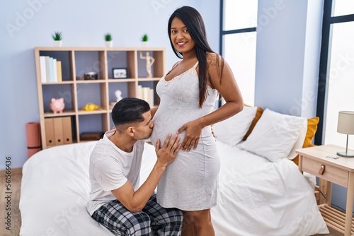 Young latin couple expecting baby kissing tummy at bedroom