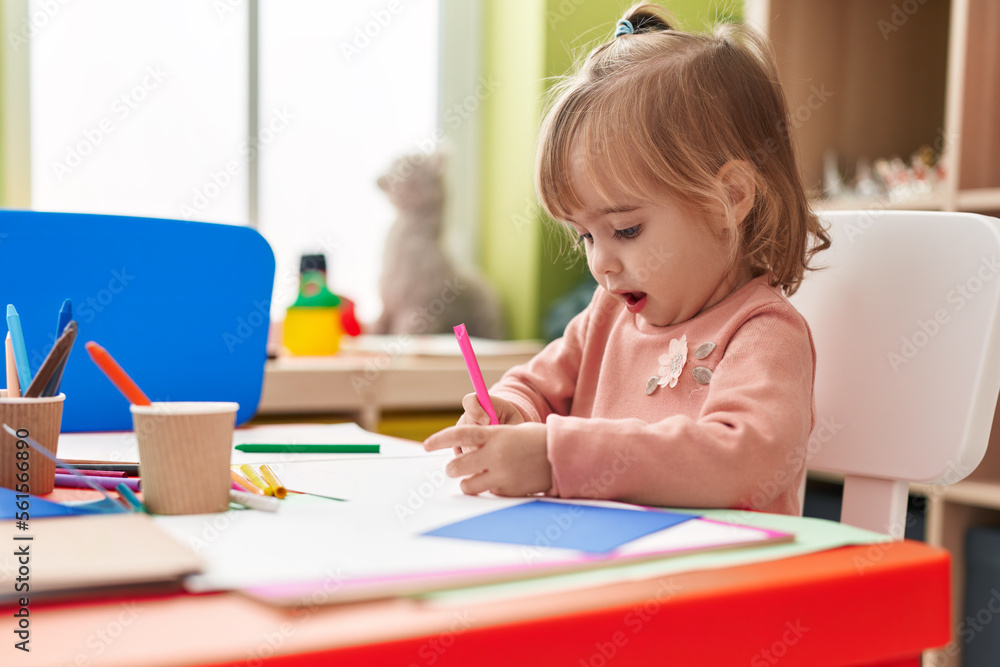 Adorable blonde girl preschool student sitting on table drawing on paper at kindergarten Adorable blonde girl preschool student sitting on table drawing on paper at kindergarten