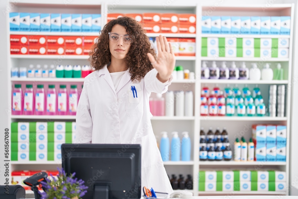 Hispanic woman with curly hair working at pharmacy drugstore doing stop sing with palm of the hand. warning expression with negative and serious gesture on the face.