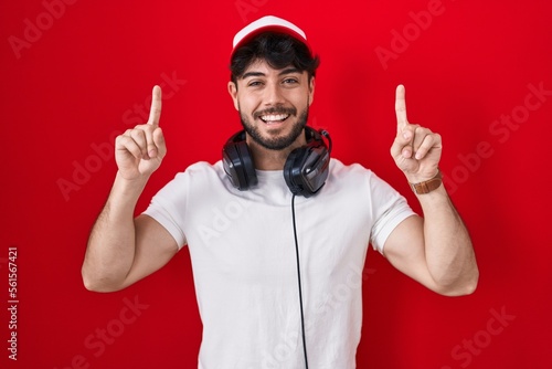 Hispanic man with beard wearing gamer hat and headphones smiling amazed and surprised and pointing up with fingers and raised arms.