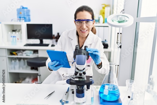 Young hispanic woman wearing scientist uniform using touchpad and microscope at laboratory