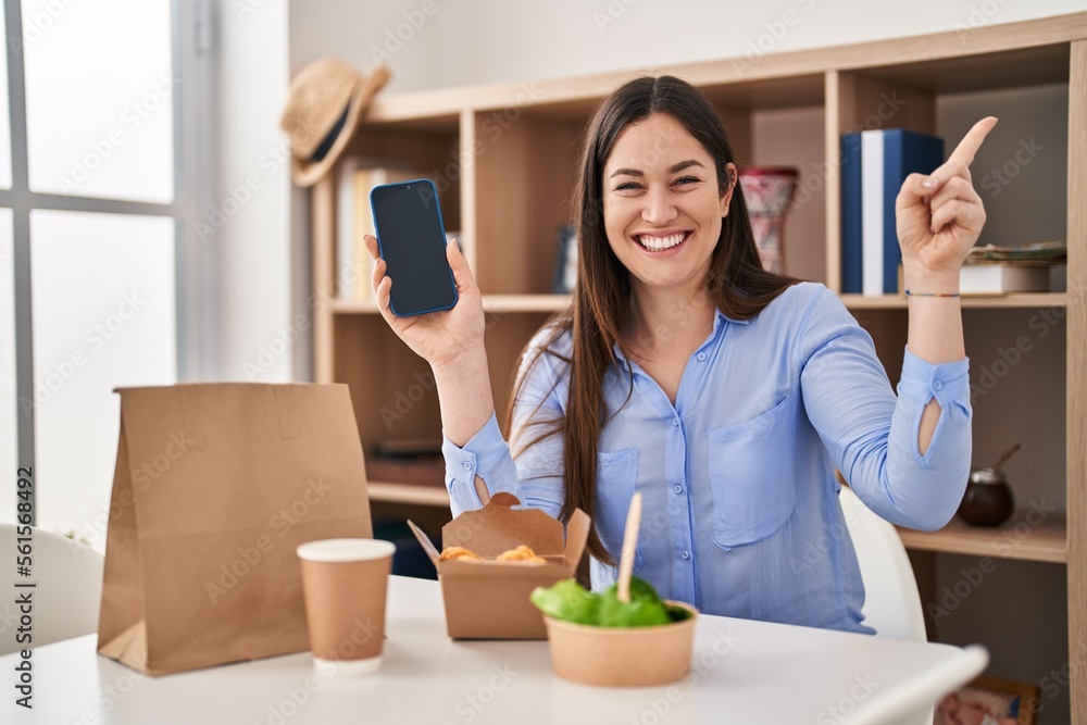 Young brunette woman eating take away food at home showing smartphone screen smiling happy pointing with hand and finger to the side Young brunette woman eating take away food at home showing smartphone screen smiling happy pointing with hand and finger to the side