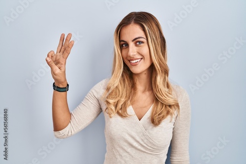 Young blonde woman standing over isolated background showing and pointing up with fingers number three while smiling confident and happy.
