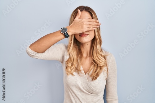 Young blonde woman standing over isolated background covering eyes with hand, looking serious and sad. sightless, hiding and rejection concept