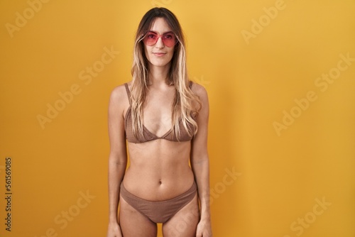 Young hispanic woman wearing bikini over yellow background relaxed with serious expression on face. simple and natural looking at the camera.
