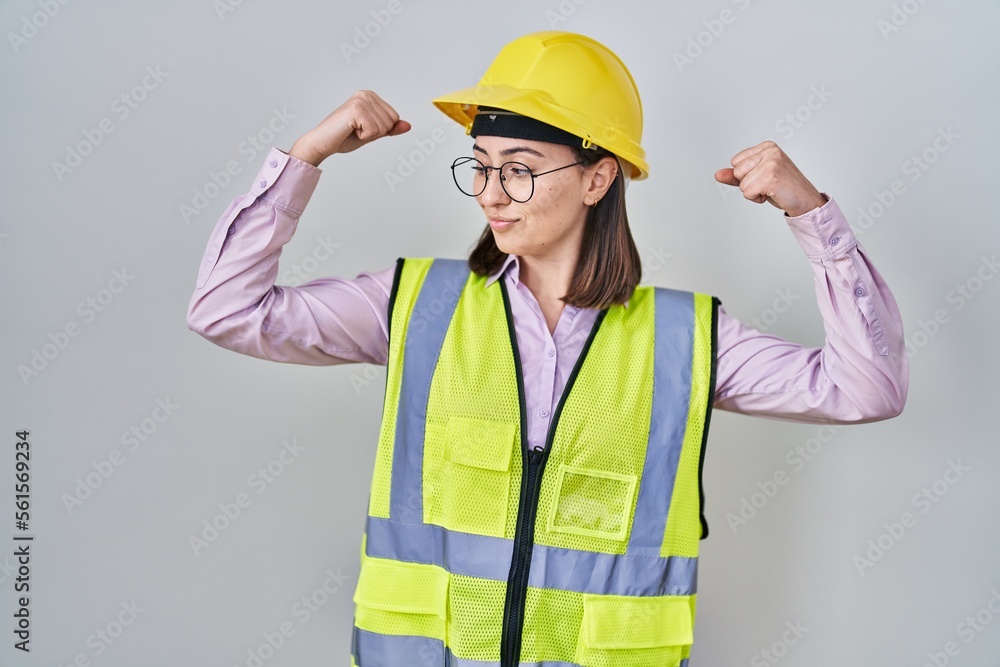 Hispanic girl wearing builder uniform and hardhat showing arms muscles smiling proud. fitness concept. Hispanic girl wearing builder uniform and hardhat showing arms muscles smiling proud. fitness concept.