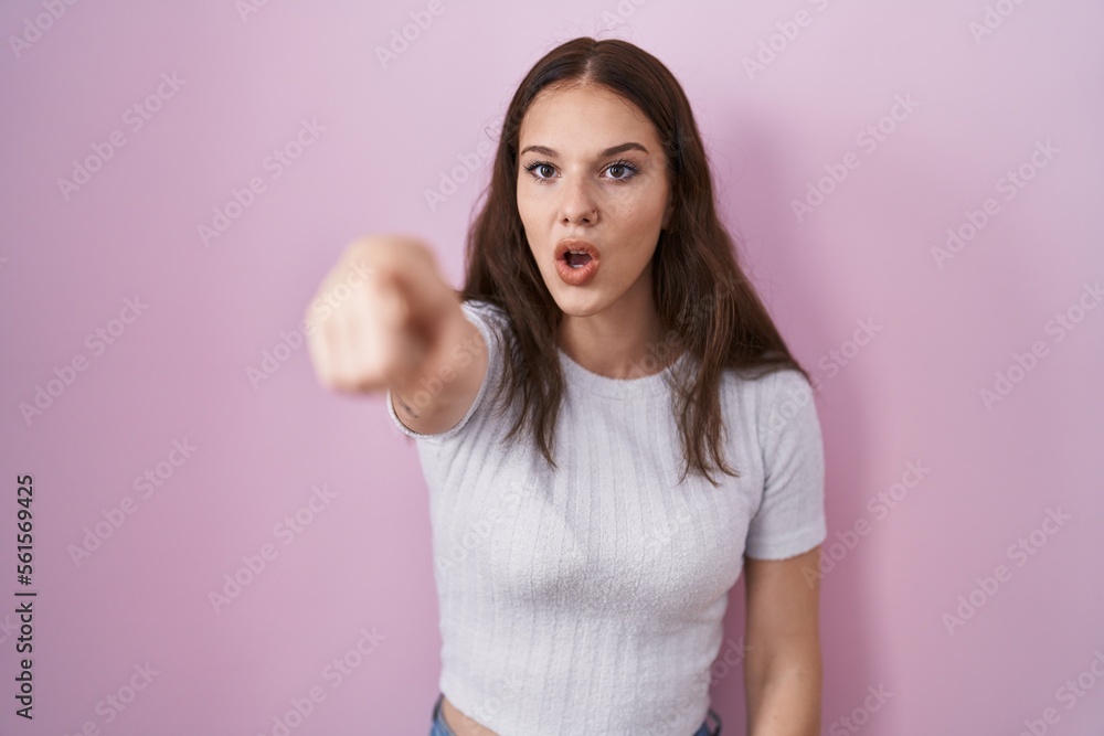 Fototapeta premium Young hispanic girl standing over pink background pointing with finger surprised ahead, open mouth amazed expression, something on the front