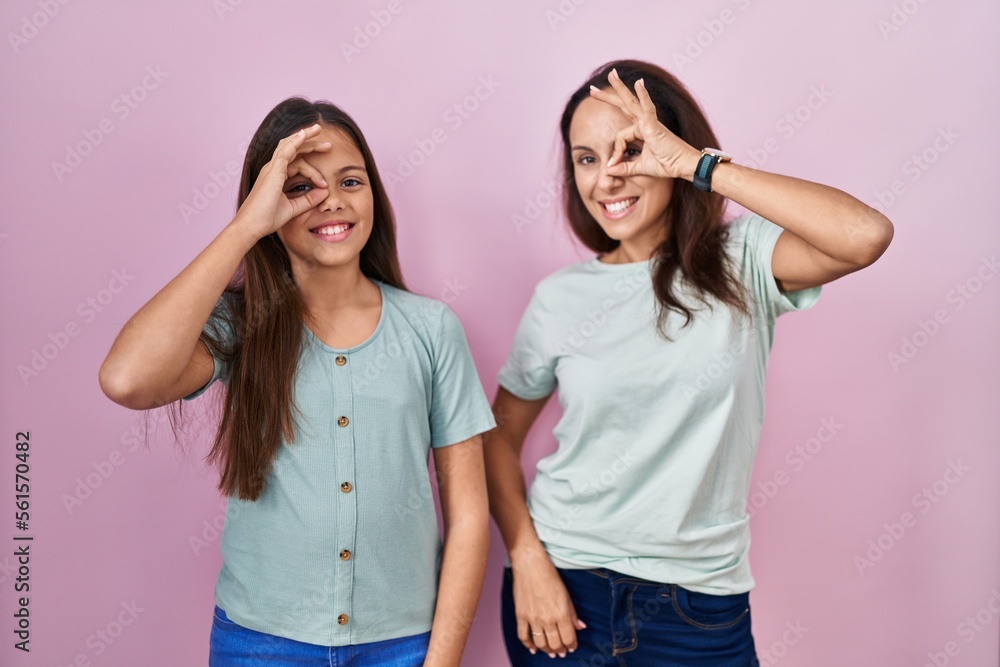 Young mother and daughter standing over pink background doing ok gesture with hand smiling, eye looking through fingers with happy face. Young mother and daughter standing over pink background doing ok gesture with hand smiling, eye looking through fingers with happy face.