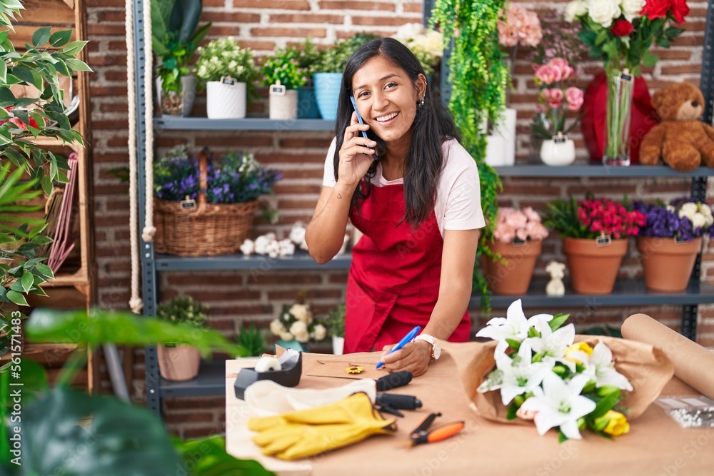 Young beautiful hispanic woman florist talking on smartphone writing on envelope letter at flower shop