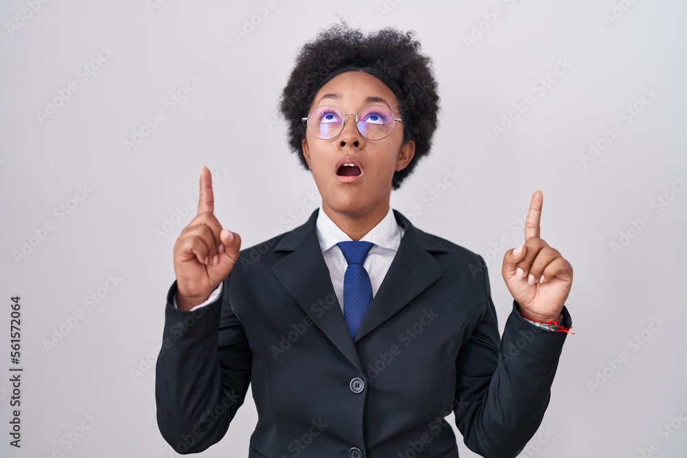 Beautiful african woman with curly hair wearing business jacket and glasses amazed and surprised looking up and pointing with fingers and raised arms. Beautiful african woman with curly hair wearing business jacket and glasses amazed and surprised looking up and pointing with fingers and raised arms.