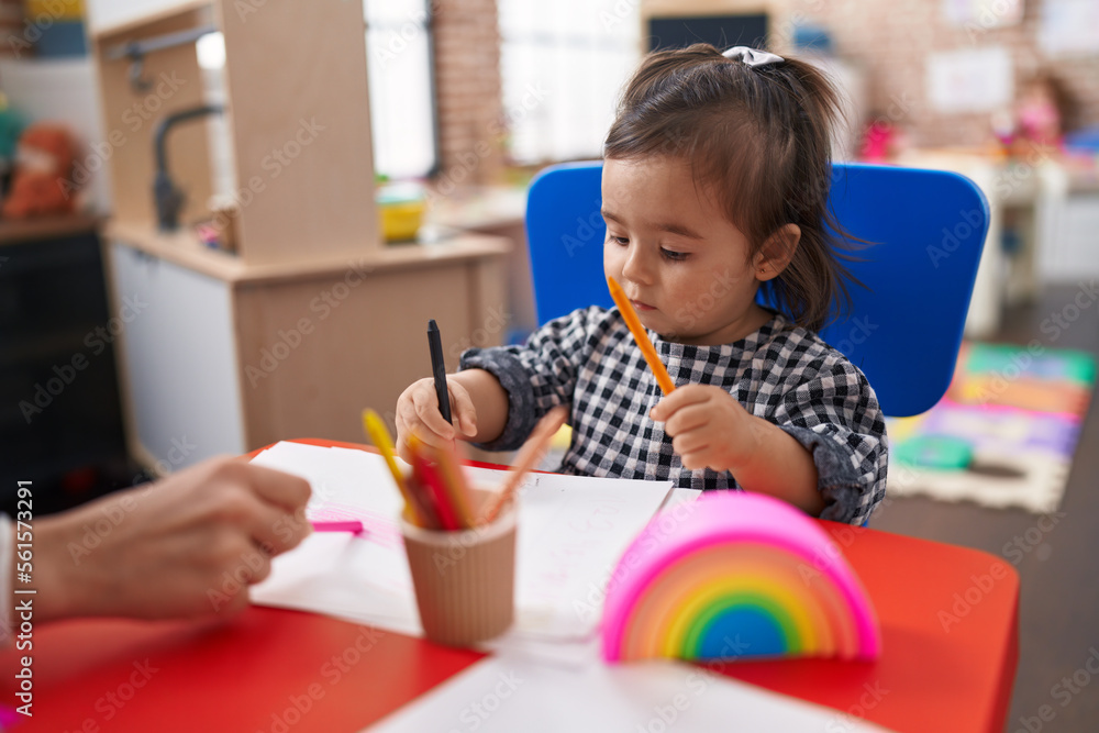 Adorable chinese girl preschool student sitting on table drawing on paper at kindergarten Adorable chinese girl preschool student sitting on table drawing on paper at kindergarten
