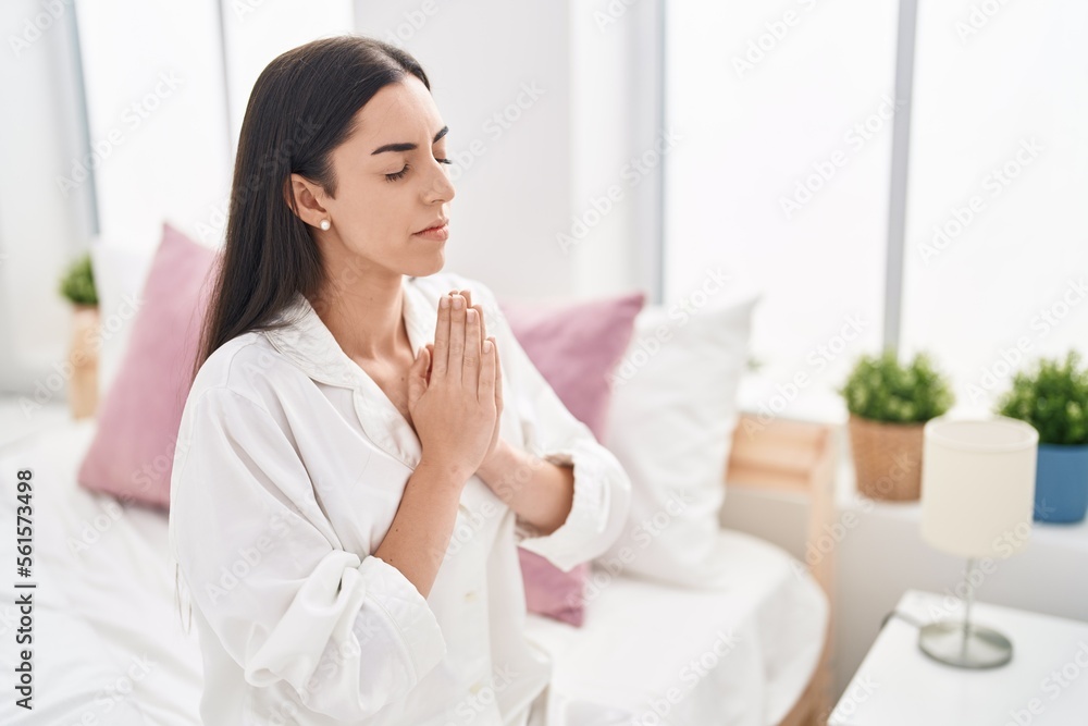 Young hispanic woman praying sitting on bed at bedroom